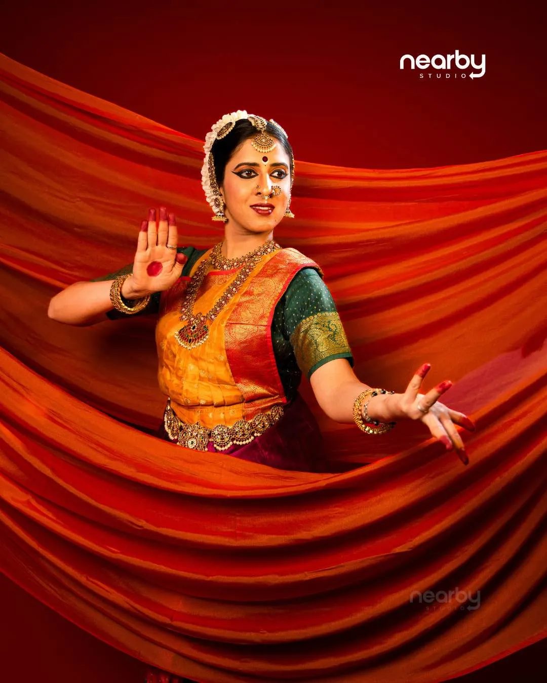 Bharatanatyam dancer in a silk costume and temple jewellery holding a pose against flowing orange fabric at Nearby Studio